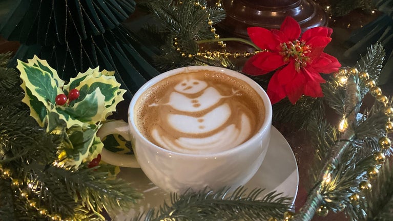 A hot milky drink in a cup on a saucer with a snowman in the foam, surrounded by Christmas decorations
